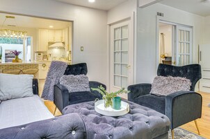 Una sala moderna con un otoman tapizado, dos sillones oscuros, una mesita auxiliar con una bandeja y vista al área de la cocina.