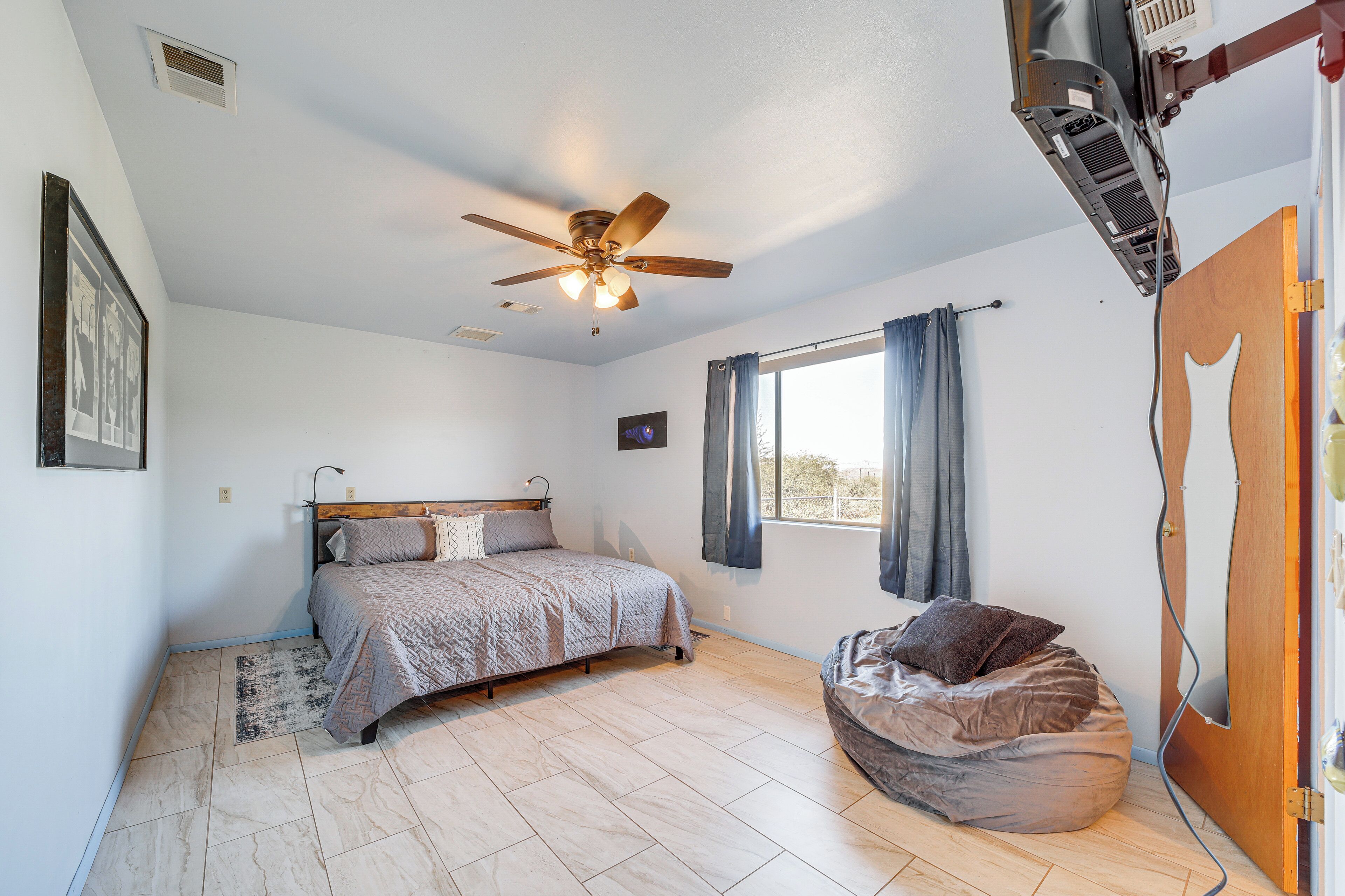 Un dormitorio con cama, ventilador de techo, ventana con cortinas y un sillón de peluche.