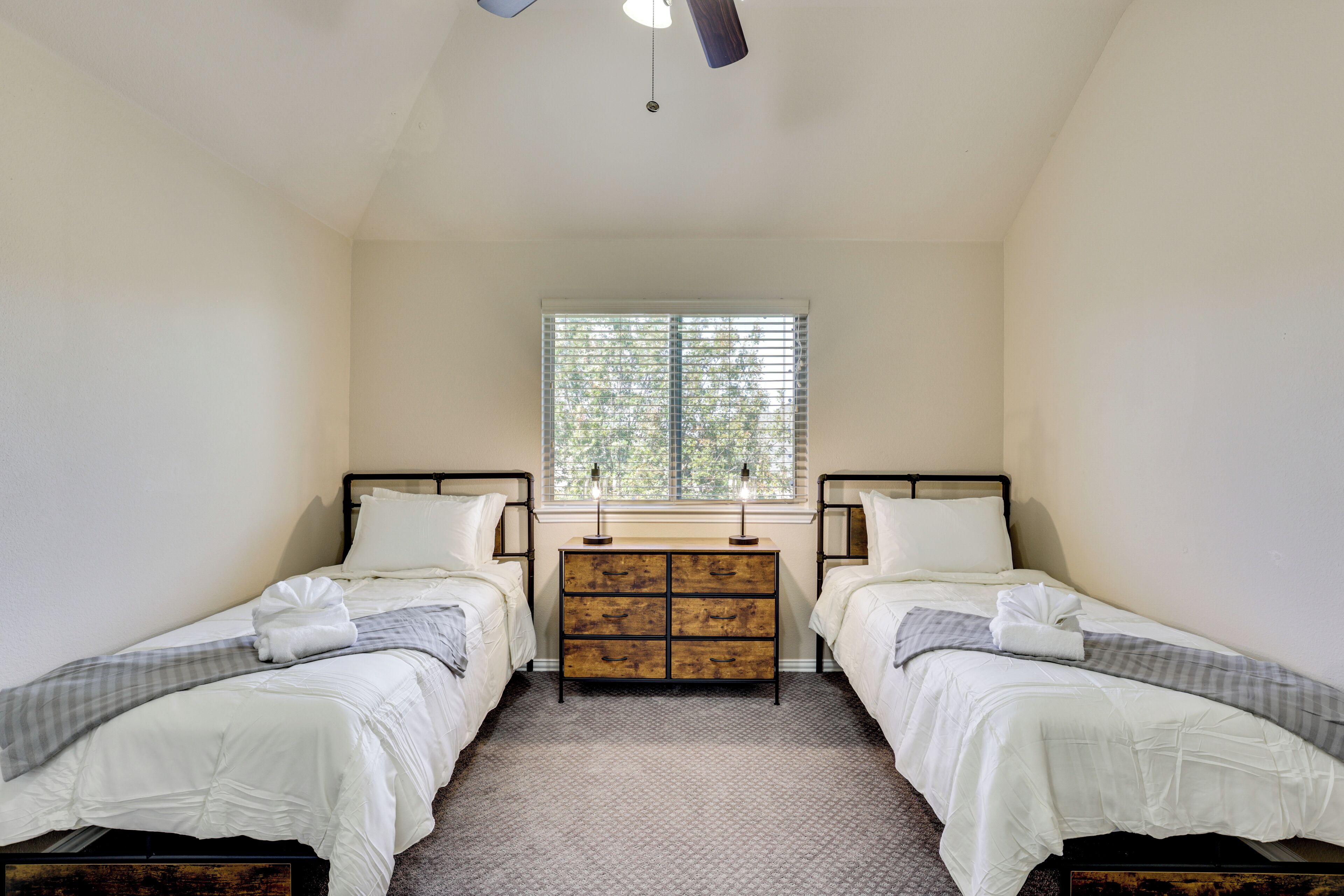 A bedroom with two single beds, a wooden dresser, a window with blinds, and a ceiling fan.