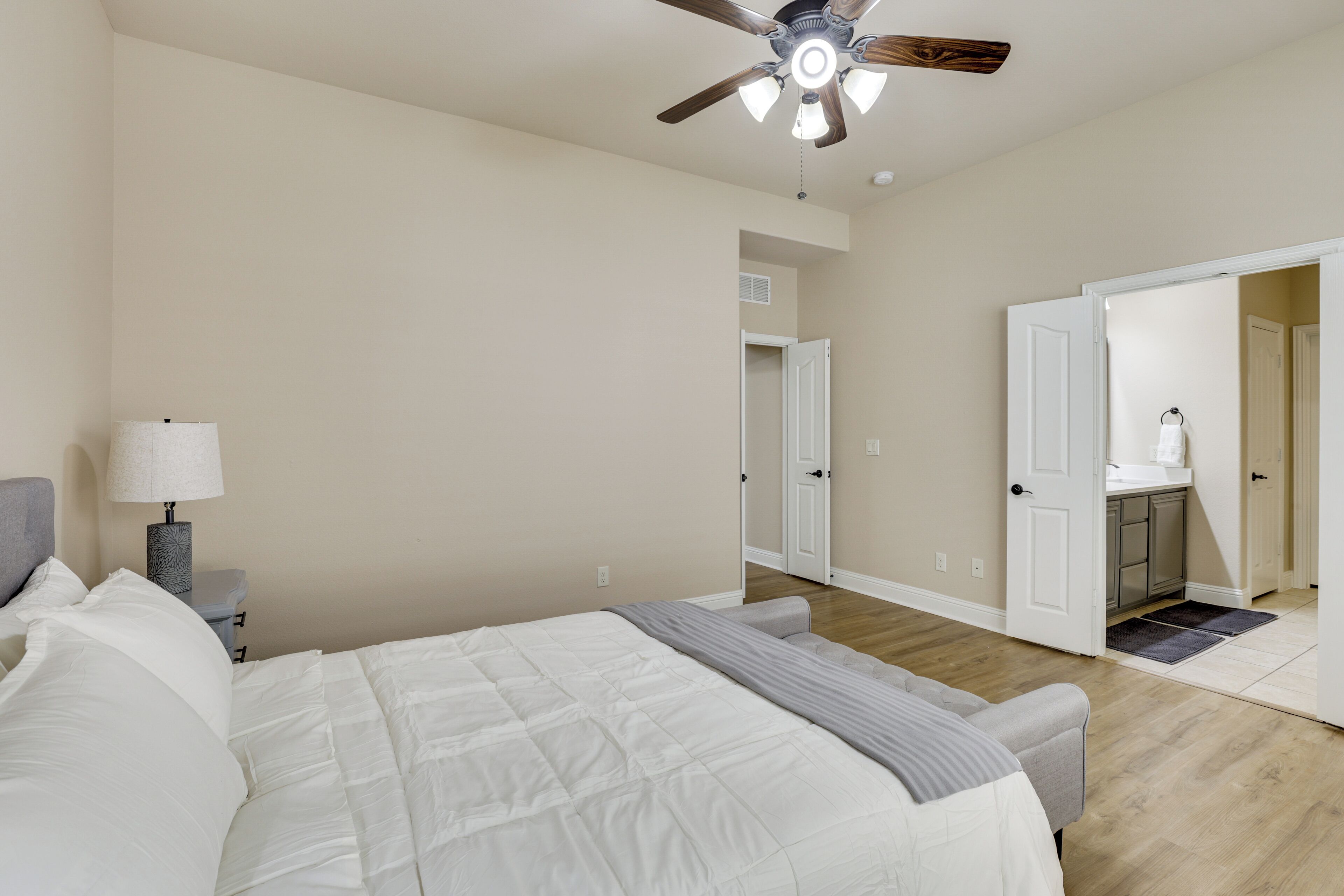 A bedroom with a bed, a nightstand, a lamp, a ceiling fan, and an open door leading to a bathroom.