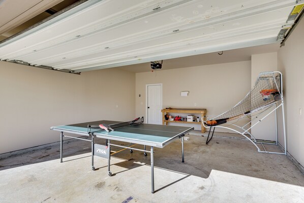 Une table de ping-pong dans un garage, avec un panier de basket et une étagère remplie d’objets.