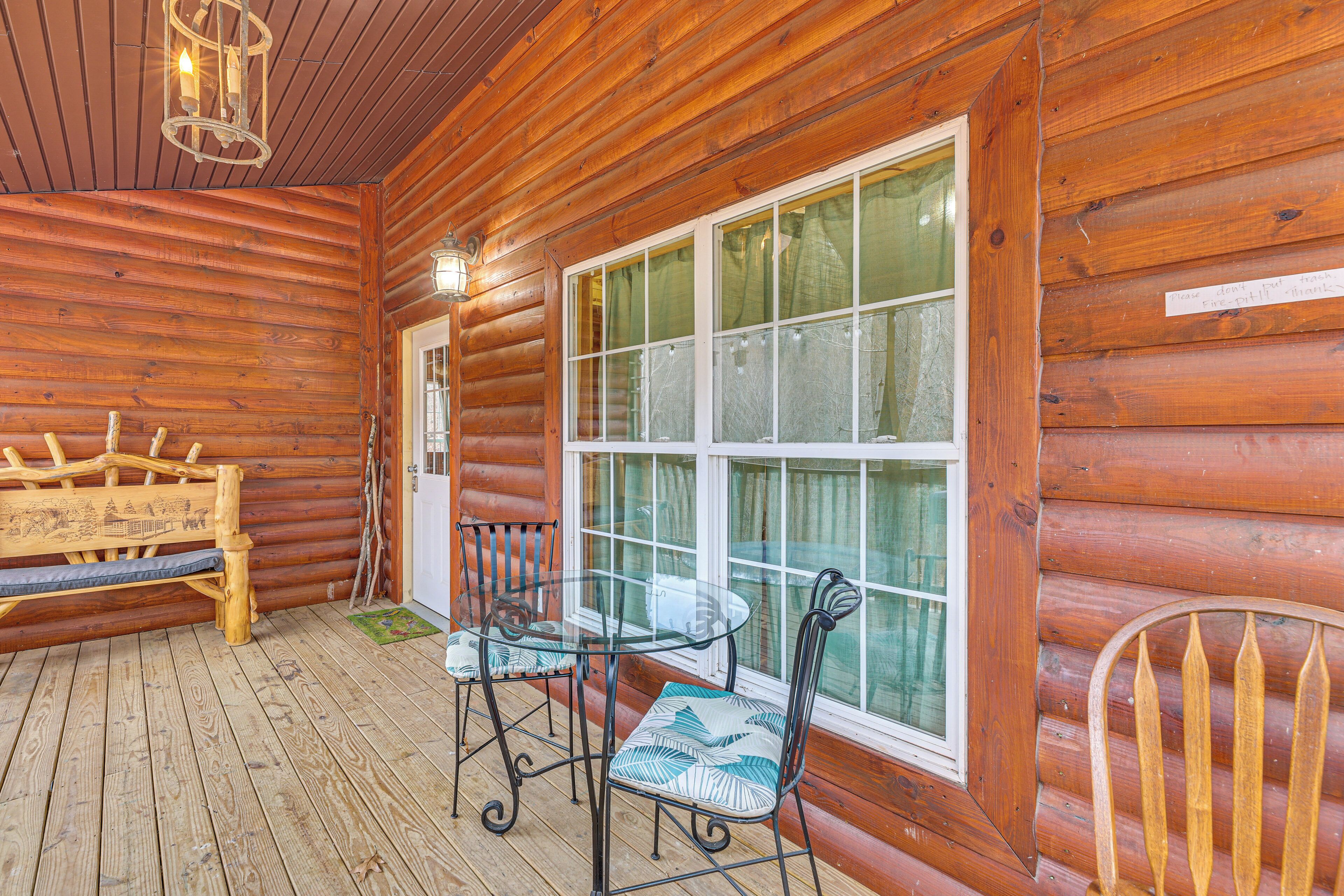 A wooden cabin with a glass table and chairs on a wooden deck, a bench, and a large window with curtains.