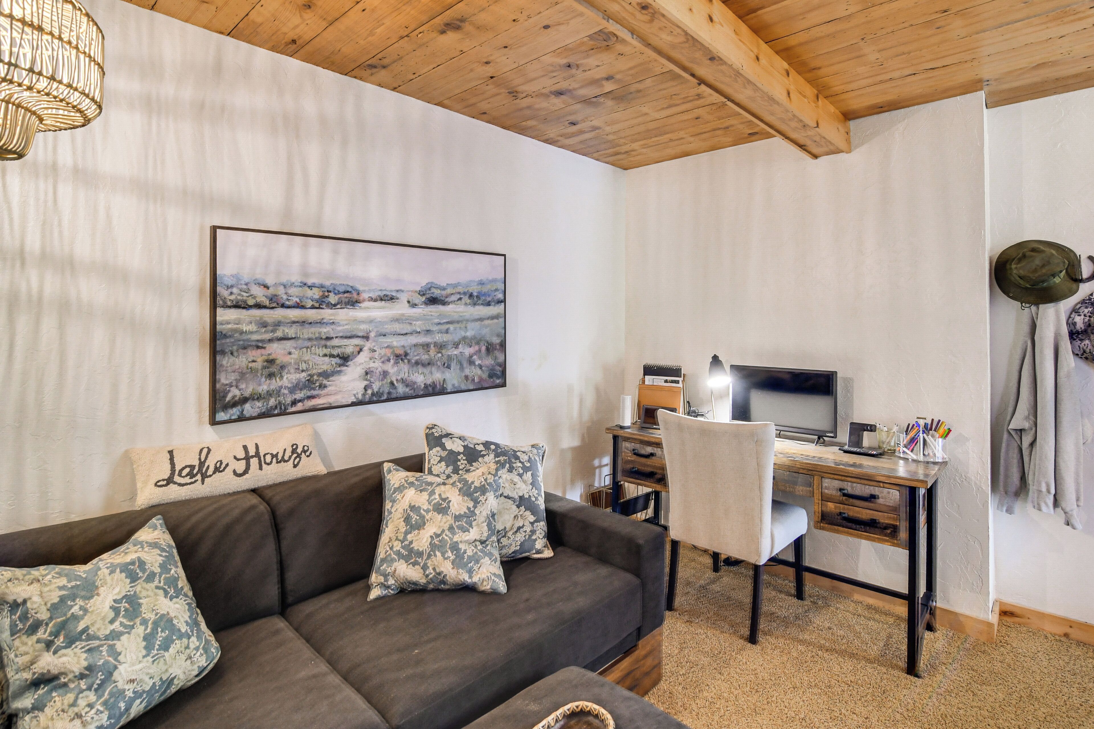 Una sala de estar con un sofá marrón oscuro, dos almohadas estampadas, un escritorio de madera con una computadora, un televisor y una lámpara fijada a la pared.