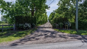 Une allée de gravier mène à une entrée fermée par un portail, le tout étant entouré d’une végétation luxuriante et de buissons en fleurs.