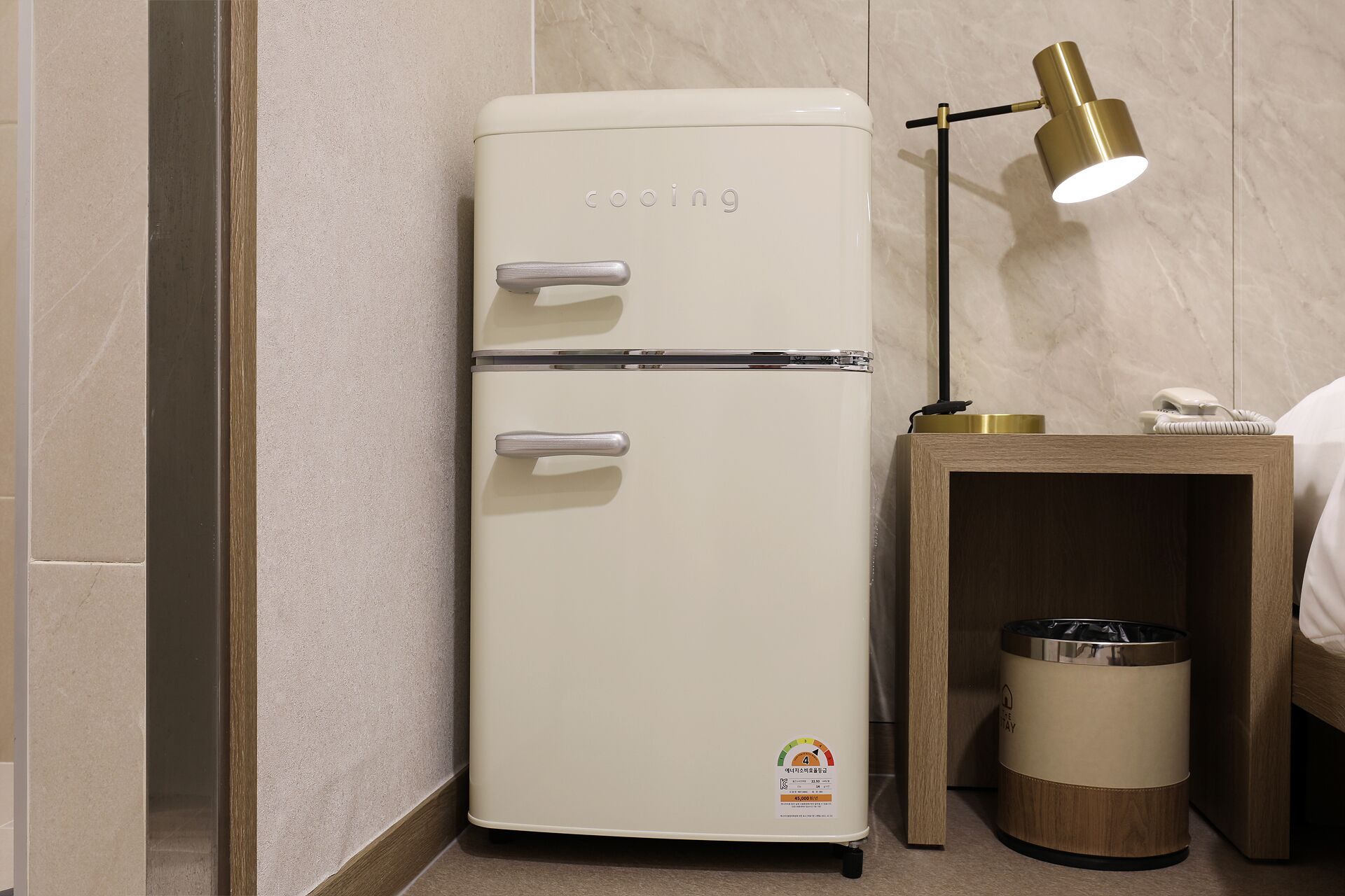 A white refrigerator with a two-door design, a small trash bin, and a wooden nightstand with a lamp.