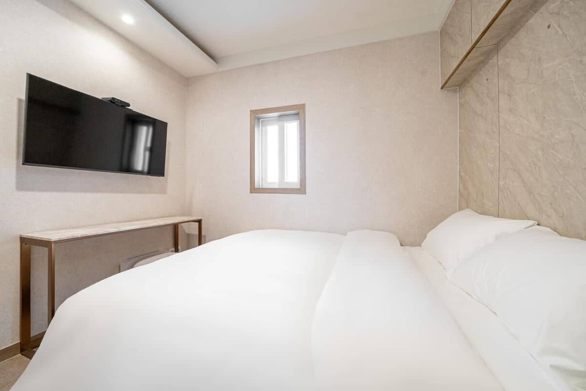 Un dormitorio con cama, televisión montada en la pared y un ventanal pequeño.