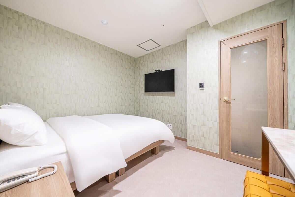 Una cama individual con estructura de madera, una mesita de noche con teléfono, un televisor montado en la pared y una puerta de madera.