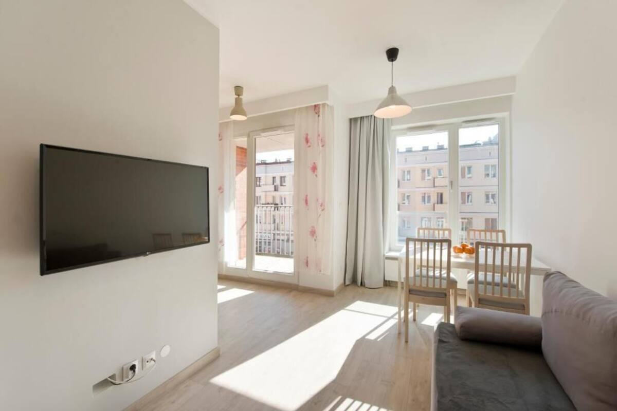 Ett modernt vardagsrum med en platt-TV, en soffa, ett matbord med stolar och stora fönster med utsikt över byggnader.