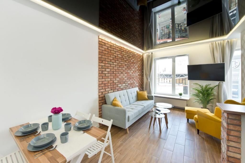 Ein modernes Wohnzimmer mit Ziegelwand, grauem Sofa, gelben Sesseln, einem Esstisch gedeckt mit Tellern und Tassen sowie einem an der Wand befestigten Fernseher.