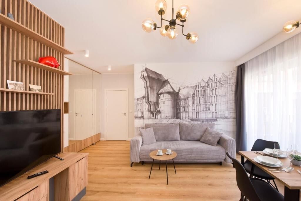 A modern living room with a flat-screen TV, wooden shelving, a grey sofa, a dining table with chairs, and a large cityscape mural on the wall.