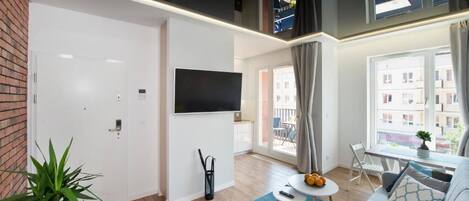 A modern living room with a flat-screen TV, a round coffee table with fruits, a grey sofa, and a large window with a view of buildings.