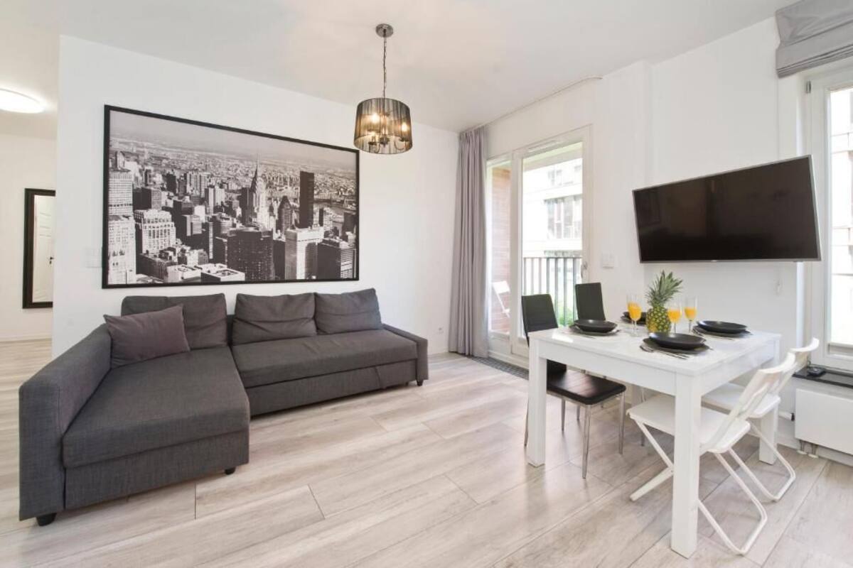 Ett modernt vardagsrum med en grå hörnsoffa, ett vitt matbord med stolar, en svart platt-TV och en stor svartvit målning som föreställer en stadsbild på väggen.