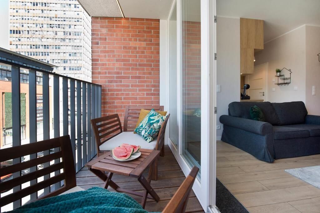 Un balcon avec un mur en briques, une table en bois sur laquelle se trouve une assiette de pastèque, et un canapé bleu à l’intérieur d’une pièce.