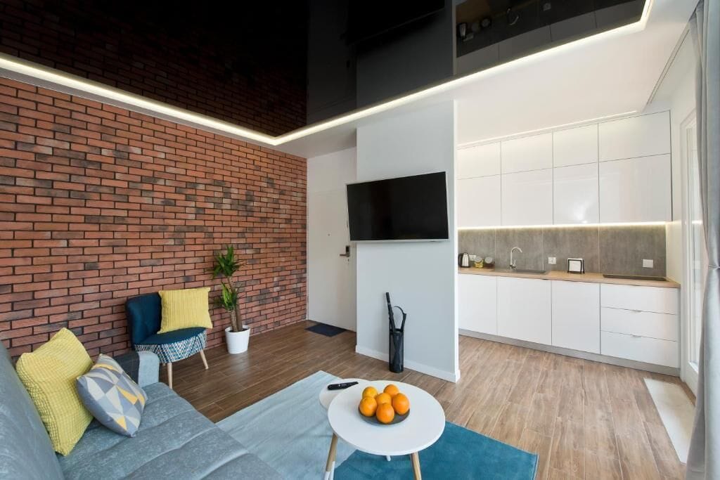 Una sala de estar moderna con pared de ladrillo, un sofá con cojines, una mesita baja con naranjas, y una zona de cocina con televisor y armarios.