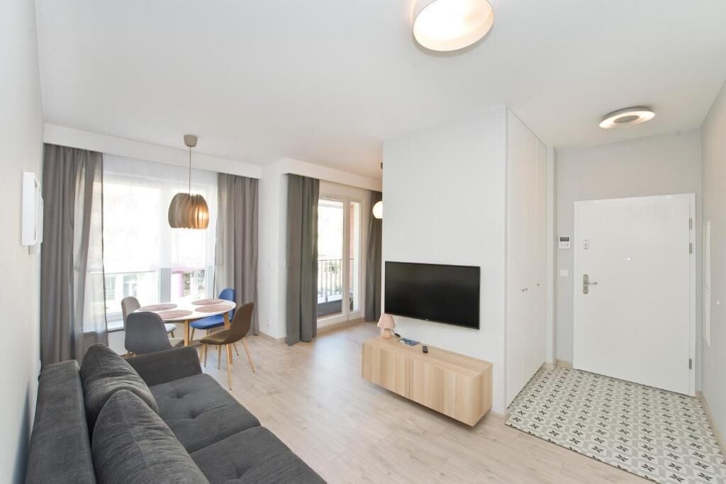 A modern living room with a sofa, a TV mounted on the wall, a dining area with a table and chairs, and a balcony with a view.