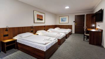 A hotel room with four single beds, a desk with a chair, a TV mounted on the wall, and two framed pictures.