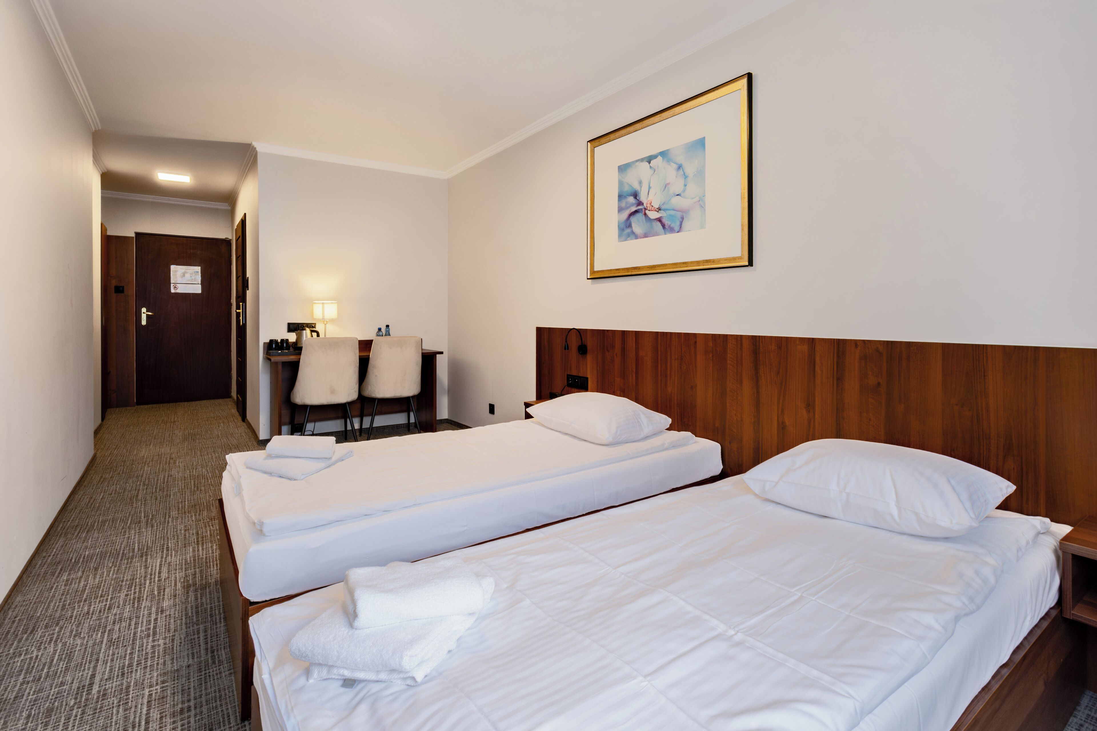 Camera d'albergo con due letti singoli, una testiera in legno, una scrivania con una sedia, una lampada e un quadro incorniciato appeso al muro.