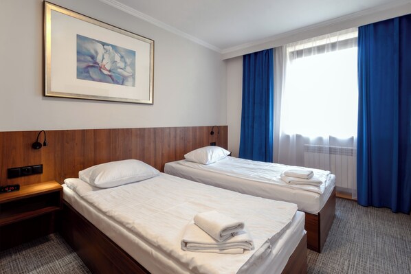 A hotel room with two beds, a wooden headboard, a framed picture on the wall, and a window with blue curtains. - Noclegi Hit Rooms & Apartments (Warsaw)