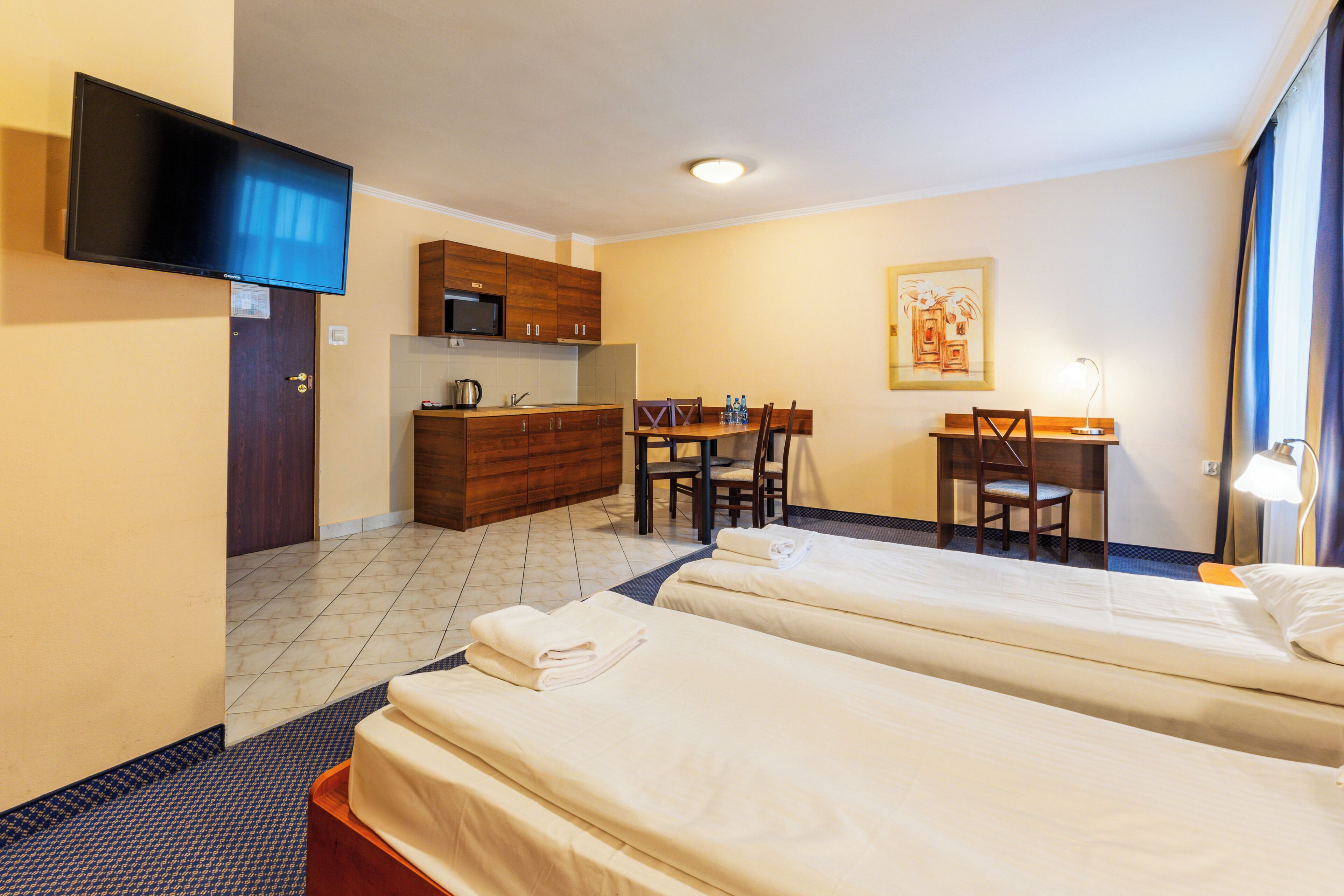 A hotel room with two beds, a desk, a chair, a television, and a kitchenette.