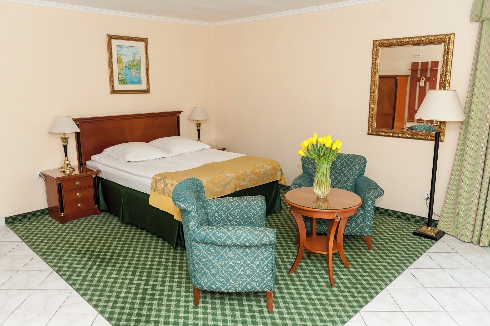 A hotel room with a bed, two bedside tables with lamps, a chair, a small round table with a vase of flowers, and a mirror on the wall.