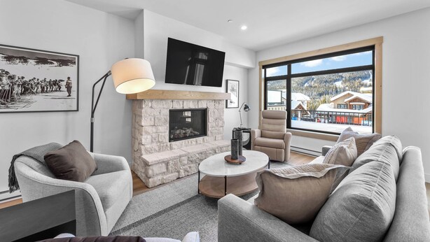 A modern living room with a stone fireplace, a large TV, and a view of a snowy landscape through a large window. - Switchback Creek Townhouse 17 (Sun Peaks)