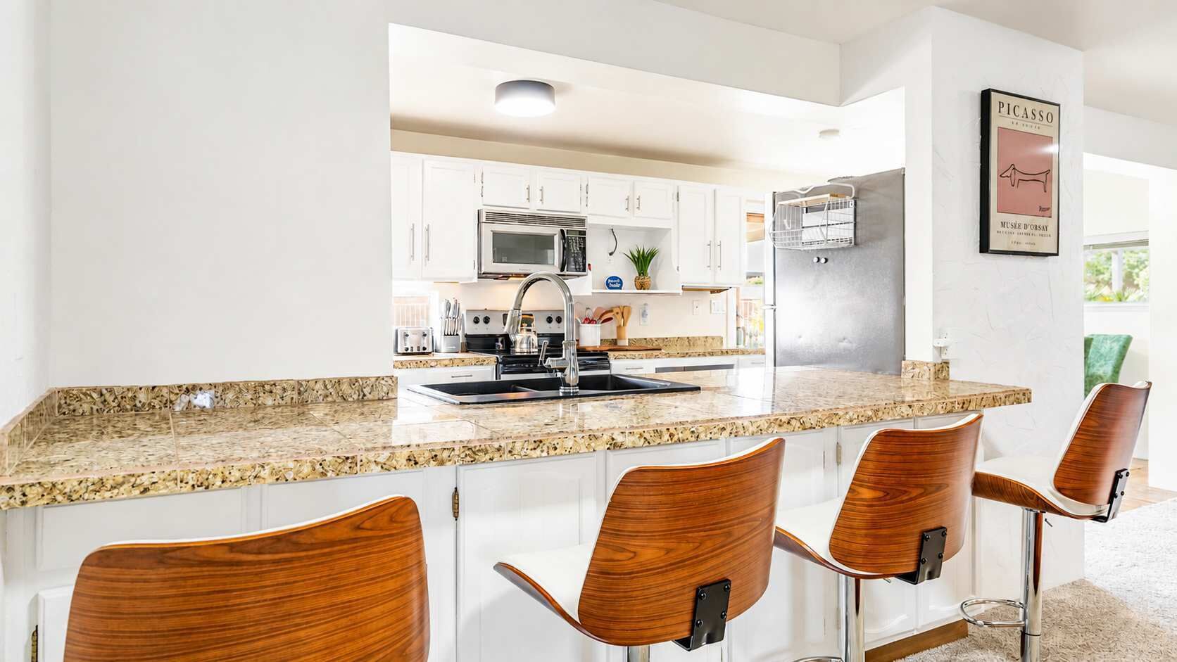 A kitchen with white cabinets, a granite countertop, a stainless steel sink, and a refrigerator. There are four wooden chairs with metal legs.