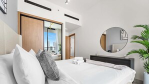 A modern hotel room with a large bed, a round mirror, a potted plant, and a view of the city through a sliding door. - StayShort - Sophisticated JVC Apartment Sleeps 4 with Balcony (JVC)