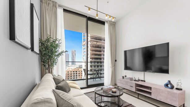 A modern living room with a sofa, a coffee table, a TV, and a balcony with a view of buildings. - StayShort - Sophisticated JVC Apartment Sleeps 4 with Balcony (JVC)