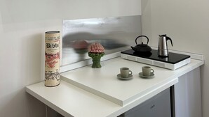 Ett kök med en vit bänkskiva, en svart vattenkokare, en liten vas med rosa blommor och en hylla med dekorativa burkar.