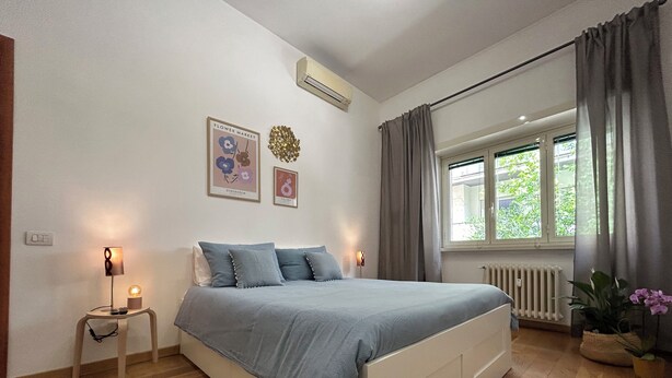 Ett sovrum med en säng, sänglampor, ett fönster med gardiner, ett element och dekorationer på väggen.