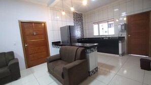 A modern kitchen with a central island, a built-in oven, and a refrigerator. There is a wooden door on the left and a window with blinds on the right. - Vacation home in Pirenópolis (Pirenópolis)