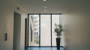 Corredor moderno de hotel com uma grande porta de vidro deslizante que dá para uma varanda, um vaso de planta e um edifício visível do lado de fora.