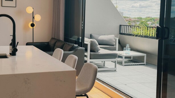 Quarto moderno com escrivaninha branca, cadeira preta, sofá cinza e mesa de centro branca. Há uma janela grande com vista para área verde e paisagem urbana.
