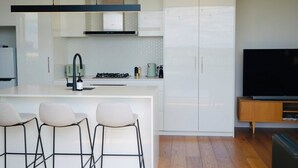 Uma cozinha moderna com armários brancos, uma ilha central com banquetas, fogão embutido e uma TV de tela plana em um suporte de madeira.