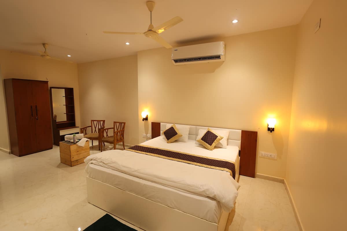 A bedroom with a bed, a ceiling fan, an air conditioning unit, a wooden wardrobe, a wooden chest of drawers, a chair, and two wall-mounted lamps.