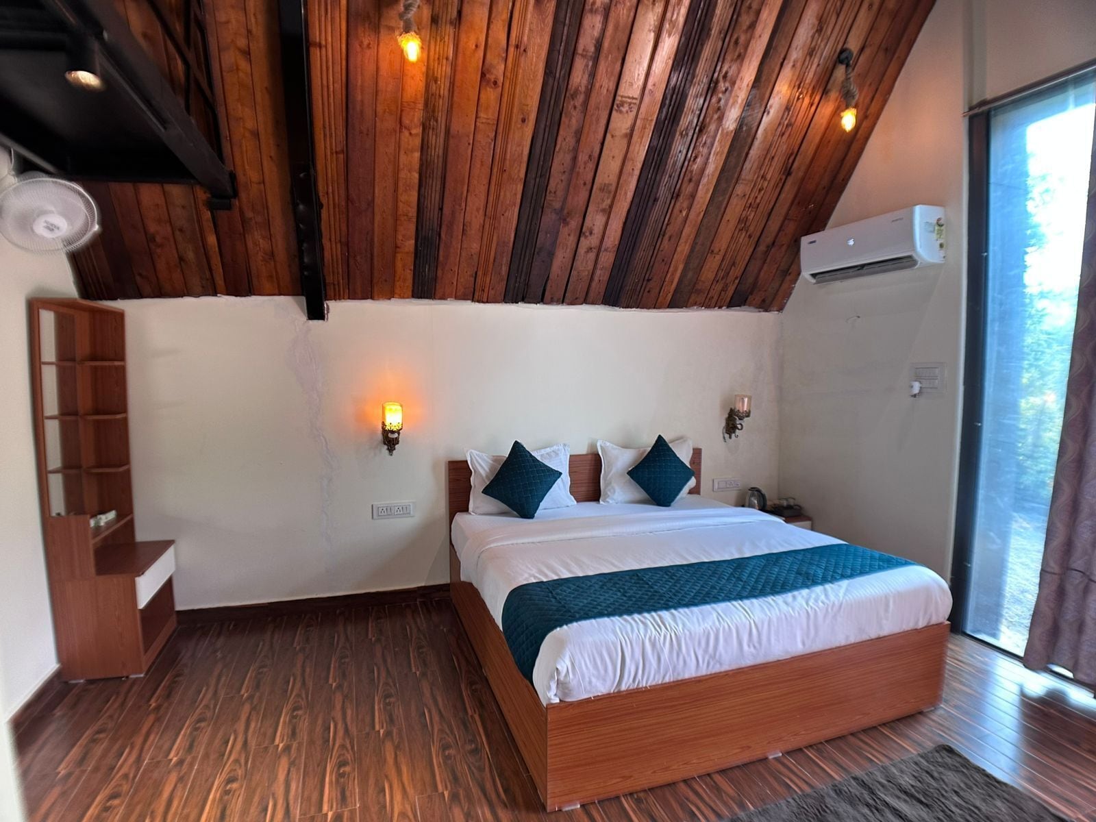 A bedroom with a wooden ceiling, a bed with white bedding and blue pillows, a wooden floor, and a window with curtains.