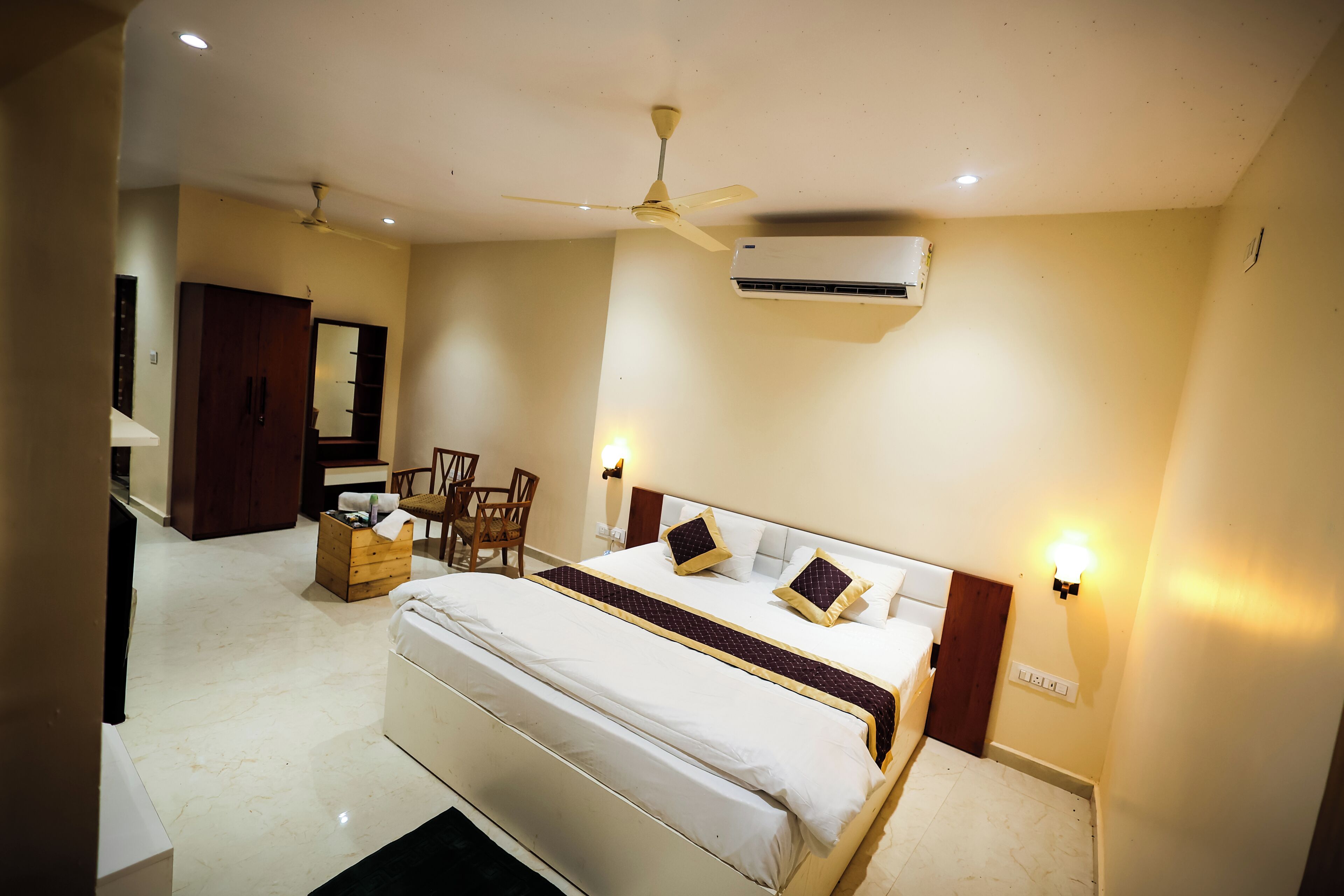 Habitación de hotel con cama, ventilador de techo, aire acondicionado, escritorio con silla y espejo.
