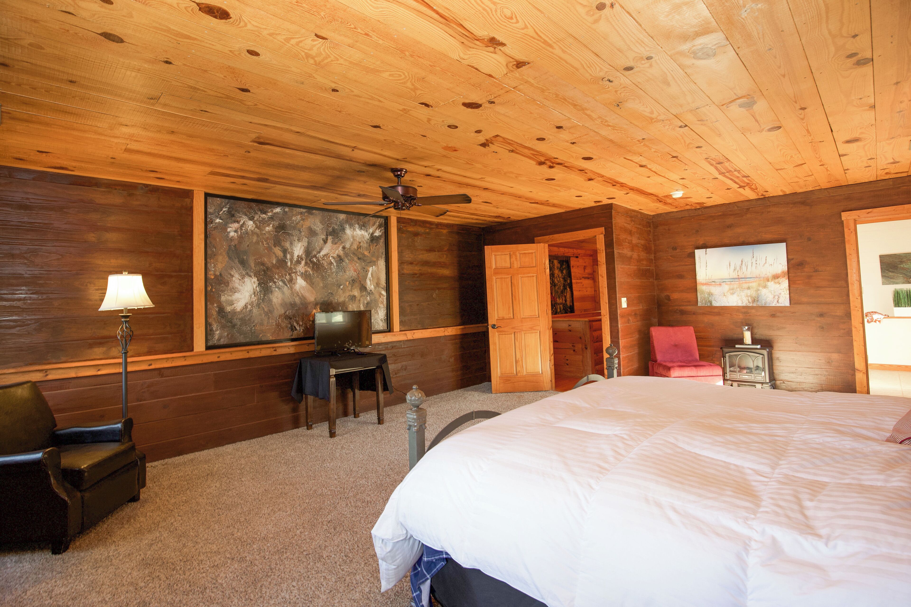Une chambre dans une cabane en bois, avec un lit, un bureau, une chaise, une lampe et un ventilateur de plafond.