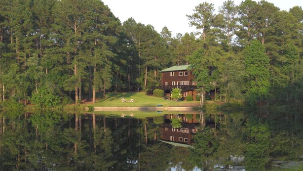 Una casa de dos pisos con techo verde, rodeada de un bosque denso y reflejada en un lago tranquilo.
