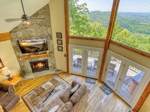 A cozy living room with a stone fireplace, a flat-screen TV, a sofa, and a large window offering a scenic view. - Peekin'Over Pigeon Forge 3 bedroom, 3 bath cabin (Sevierville)