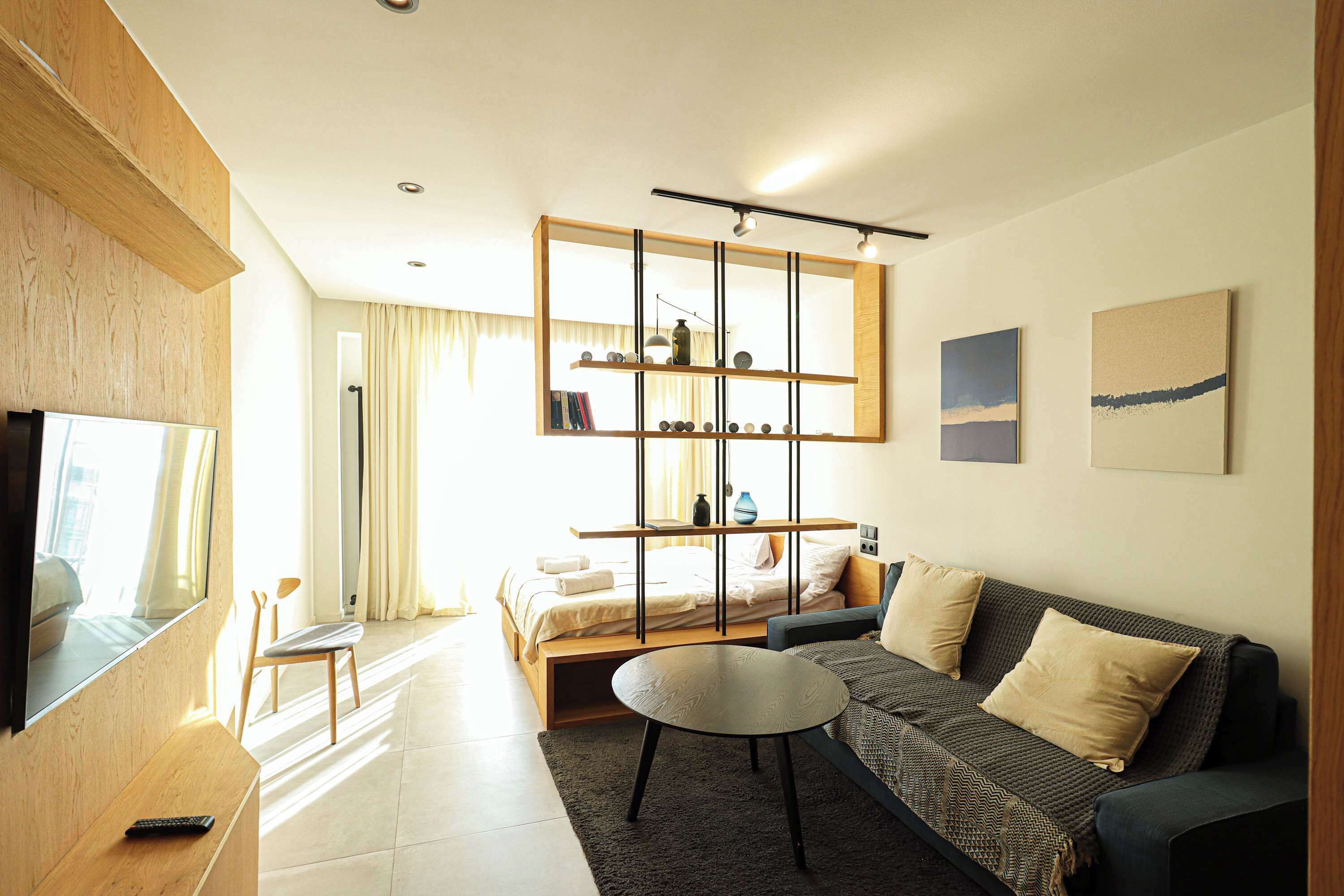 Una moderna camera d'albergo con divano, un tavolino rotondo, una TV a muro, una libreria e un letto con vista sulla città.