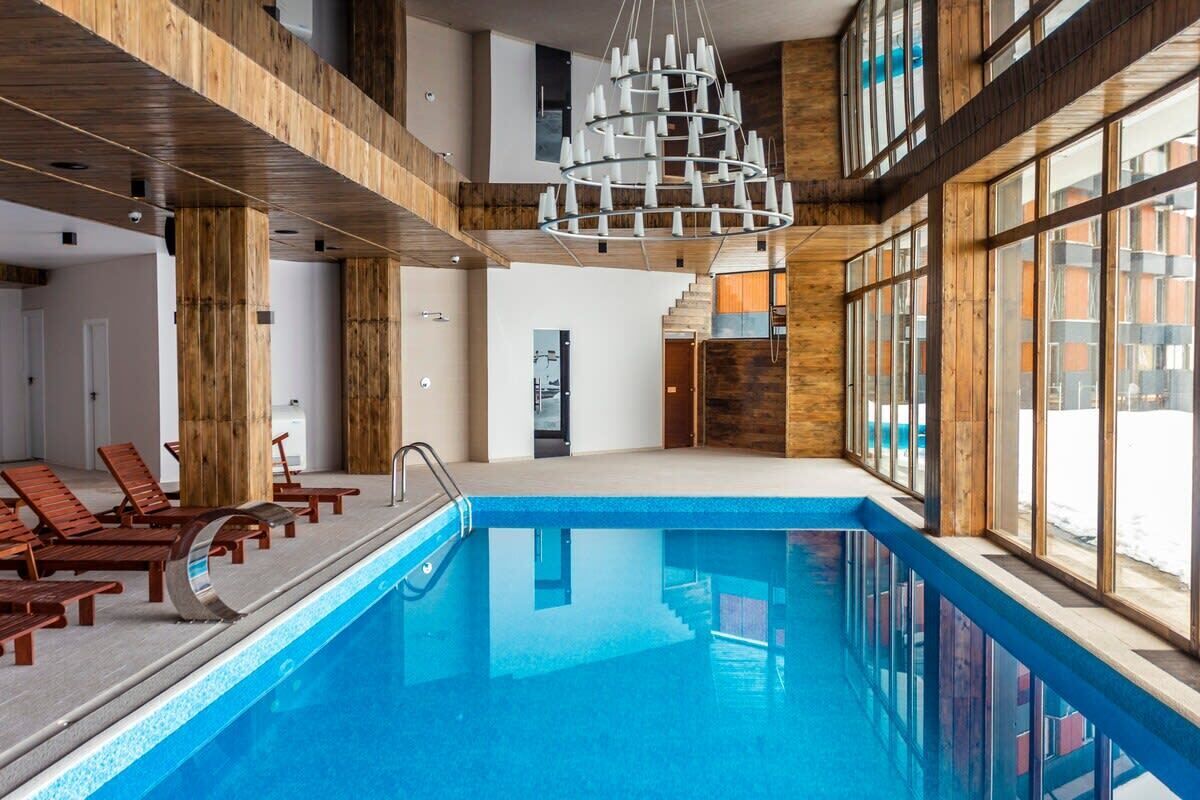 A modern indoor swimming pool with wooden beams, a central light fixture, and a view of the outside.