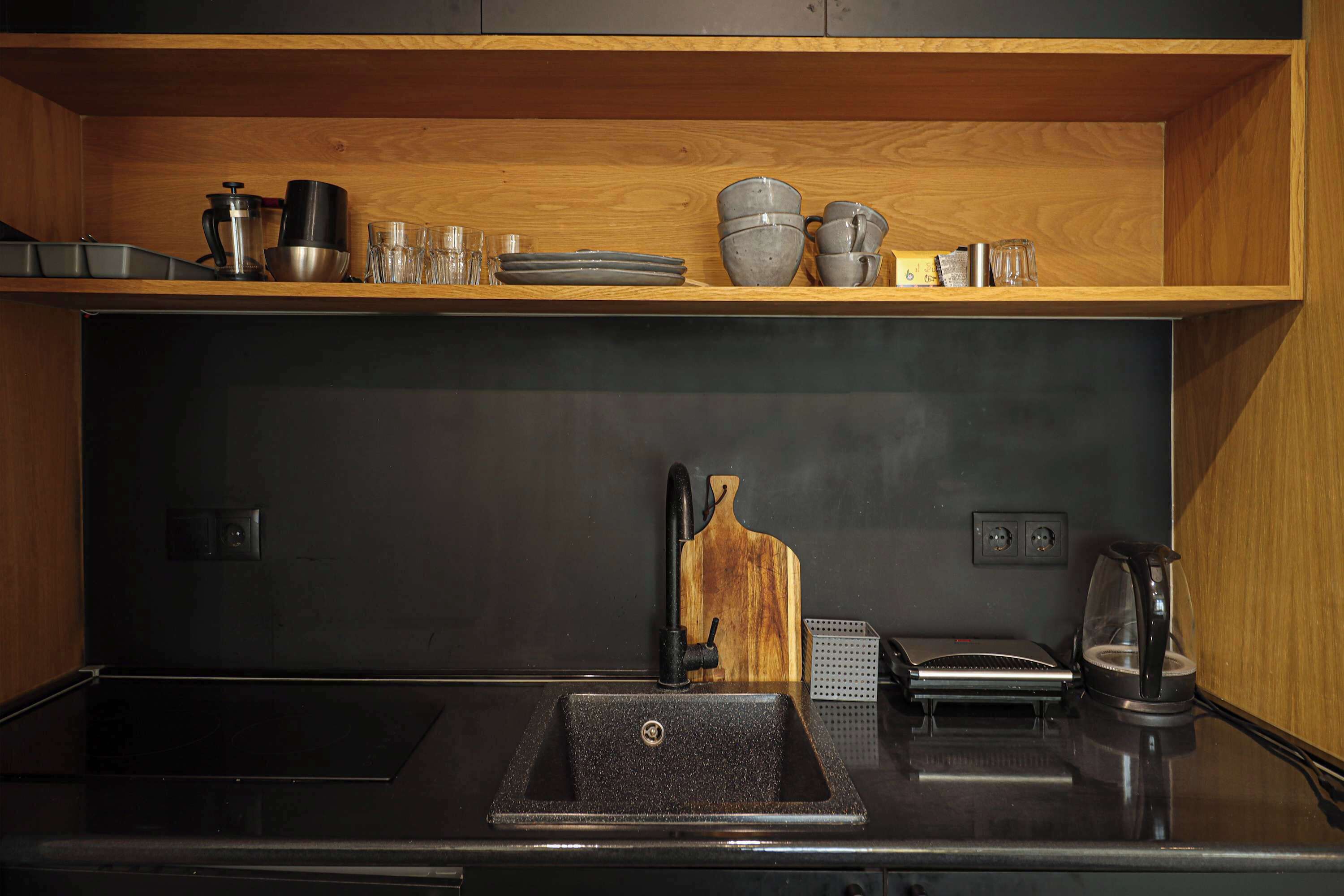Une cuisine avec un évier noir, une étagère en bois au-dessus où sont rangés divers ustensiles de cuisine, et une machine à café posée sur le plan de travail.
