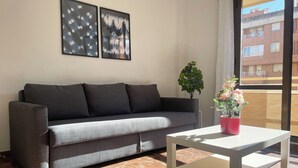 A grey sofa with cushions, a small white table with a red vase, and a potted plant. - Apartamentos el Pilar Suites 3000 (Zaragoza)