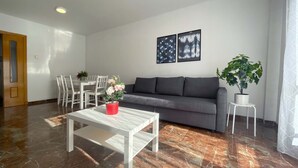 A modern living room with a grey sofa, white coffee table, and a dining area with white chairs. There are two framed pictures on the wall and a potted plant on a small table. - Apartamentos el Pilar Suites 3000 (Zaragoza)