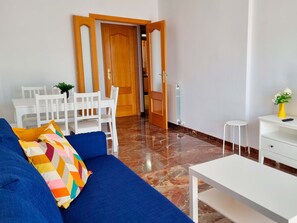 Una sala de estar con un sofá azul, una mesa de centro blanca y un comedor con una mesa y sillas blancas.