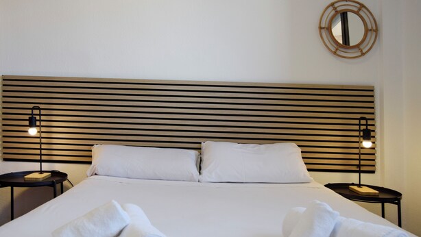 A neatly made bed with two pillows, two bedside tables with lamps, and two rolled towels on the bed. - Apartamentos el Pilar Suites 3000 (Zaragoza)