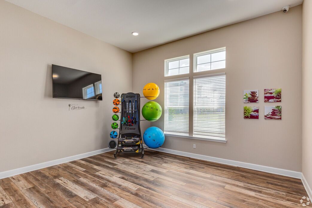 A spacious room with a wooden floor, a mounted TV, exercise equipment, and windows with blinds.