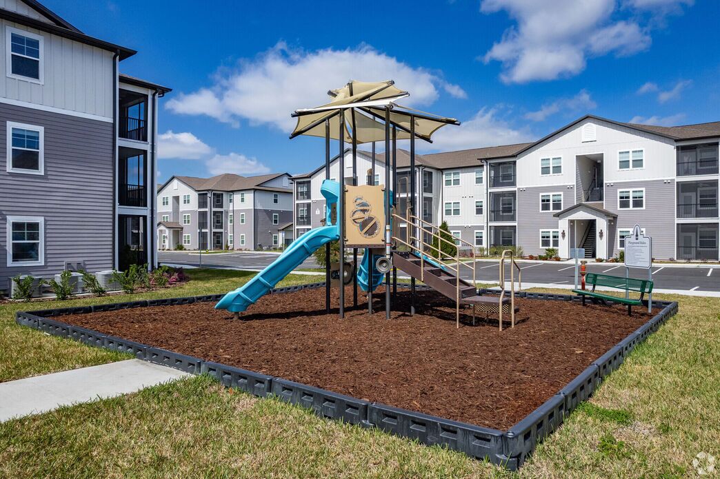 A playground with a slide and climbing structures, surrounded by a landscaped area with mulch and a bench.