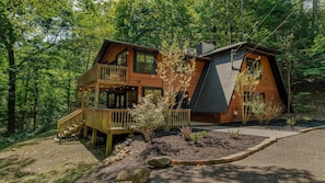 A cabin with a deck and a sloped roof, surrounded by a forest. - Sugar Bear Cabin- 4bdrm, Semi-Private, Fire-pit, Hot Tub, and Tranquil setting (Pigeon Forge)